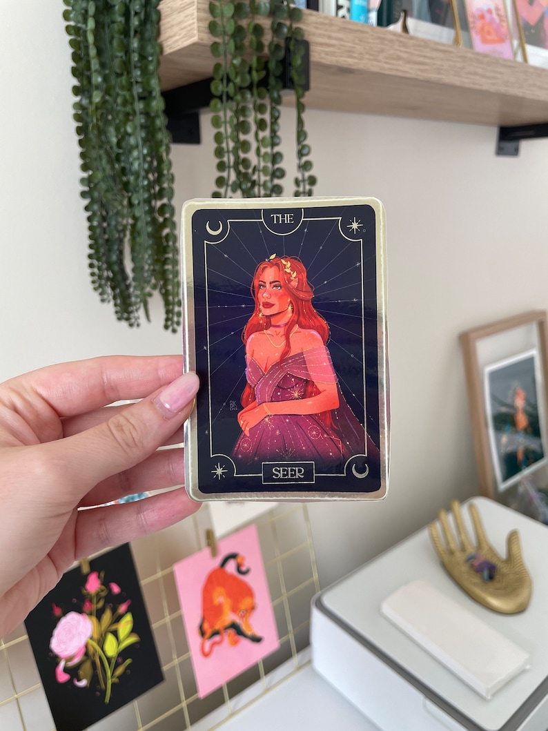 Op de afbeelding: Een tarotkaart met een gouden rand en een blauwe achtergrond. De kaart toont een vrouw met rood haar die een paarse jurk met gouden sterren draagt. De kaart heeft onderaan de tekst "THE SEER".