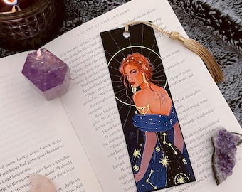 Op de afbeelding: Zwarte boekenlegger met een hemelse illustratie van een vrouw in een sterrenjurk. De boekenlegger heeft een gouden kwast en ligt op een open boek met de titel "A Court of Mist and Fury".