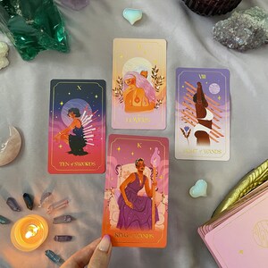 May include: Four tarot cards laid out on a purple surface with crystals and a lit candle. The cards are "Ten of Swords", "Lovers", "Eight of Wands", and "King of Wands".