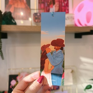May include: A rectangular paper bookmark featuring a colorful illustration of two figures wearing cowboy hats, embracing in a field at sunset. The bookmark has a light blue sky background, a blue tassel at the top, and a soft, matte finish throughout.