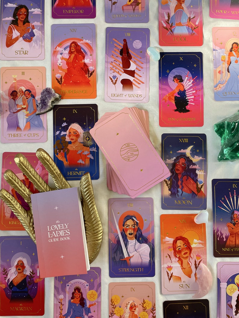 May include: A set of tarot cards with colorful illustrations of women on each card. The cards are arranged in a grid pattern on a white surface. The cards feature a variety of colors, including pink, blue, purple, and yellow. The cards are surrounded by a variety of crystals and a gold hand.