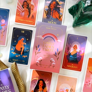 May include: A set of tarot cards with colorful illustrations of women. The cards are laid out on a white surface. The cards include The Moon, Three of Swords, Eight of Cups, The Magician, Death, and Lovers. The cards are part of a deck called "The Lovely Ladies Tarot".