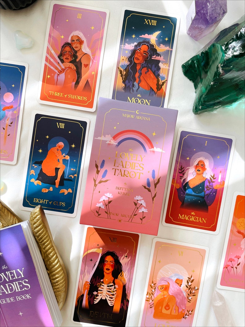 May include: A set of tarot cards with colorful illustrations of women. The cards are laid out on a white surface. The cards include The Moon, Three of Swords, Eight of Cups, The Magician, Death, and Lovers. The cards are part of a deck called "The Lovely Ladies Tarot".
