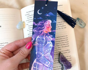 May include: A purple and blue bookmark featuring a woman in armour standing in front of a crescent moon and stars. The woman has long purple hair and is looking to the right. The bookmark has a navy blue tassel.
