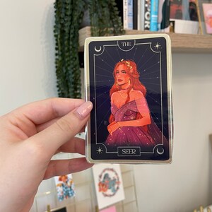 Op de afbeelding: Een tarotkaart met een gouden rand en een blauwe achtergrond. De kaart toont een vrouw met lang rood haar die een paarse jurk draagt. De kaart is getiteld "De Ziener".