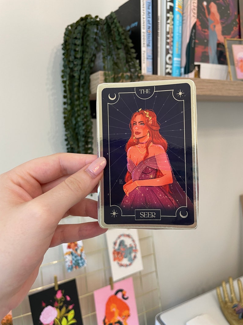 Op de afbeelding: Een tarotkaart met een gouden rand en een blauwe achtergrond. De kaart toont een vrouw met lang rood haar die een paarse jurk draagt. De kaart is getiteld "De Ziener".