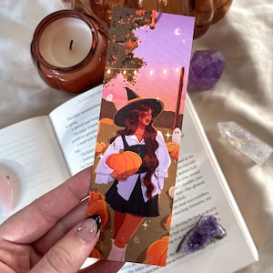 May include: A rectangular paper bookmark featuring a colorful illustration of a person with long brown hair wearing a black witch hat, a white collared shirt, and a black skirt, holding an orange pumpkin in a field with small ghosts and string lights.