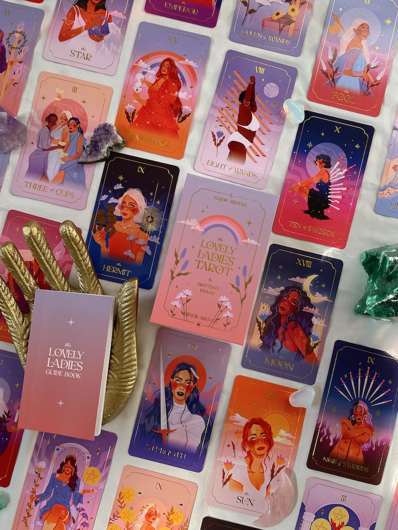 May include: A set of tarot cards with colorful illustrations of women. The cards feature a variety of scenes and symbols, including a moon, a sun, a sword, and a cup. The cards are part of a deck called "The Lovely Ladies Tarot".