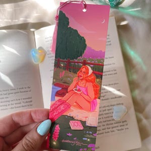 May include: A pink bookmark featuring an illustration of a person enjoying a picnic at sunset. The artwork includes a person with red hair, pink boots, and a pink dress. The bookmark has a pink tassel and the artist's signature.
