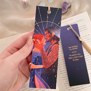 May include: A bookmark featuring a fantasy illustration of a couple embracing. The woman has red hair and is wearing a blue and red dress. The man has brown hair and is wearing a black shirt. The background is a dark blue with gold stars. The bookmark has the text "Your Power Is a Song and One I've waited a very Very Long Time To Hear" in gold lettering.