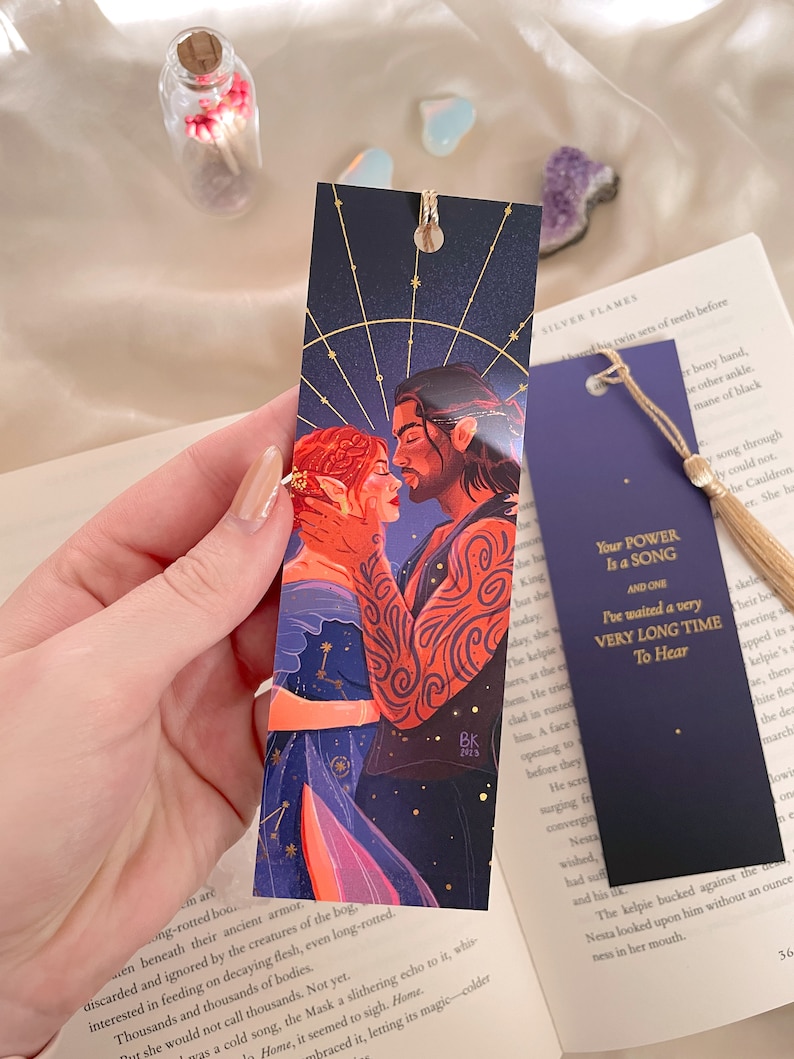 May include: A bookmark featuring a fantasy illustration of a couple embracing. The woman has red hair and is wearing a blue and red dress. The man has brown hair and is wearing a black shirt. The background is a dark blue with gold stars. The bookmark has the text "Your Power Is a Song and One I've waited a very Very Long Time To Hear" in gold lettering.