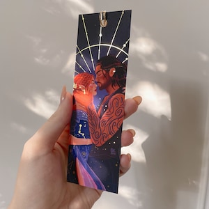 May include: A bookmark featuring a couple embracing against a dark blue background with gold stars and a gold circle around them. The woman has red hair and the man has dark hair and tattoos. The bookmark is signed "Bk 2023".