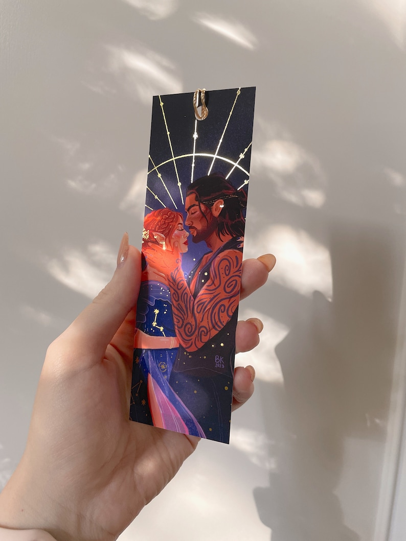 May include: A bookmark featuring a couple embracing against a dark blue background with gold stars and a gold circle around them. The woman has red hair and the man has dark hair and tattoos. The bookmark is signed "Bk 2023".