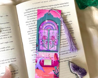 May include: A vibrant bookmark with a mermaid in a bathtub design, a window, and a purple tassel. The bookmark is placed on an open book with the title "A Court of Mist and Fury".