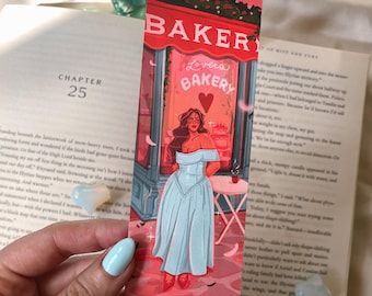 Peut inclure: Marque-page illustré d'une femme en robe bleu clair devant une boulangerie. La boulangerie a un auvent rouge avec le mot "BAKER" et "Lovers Bakery". Le marque-page est posé sur un livre ouvert.
