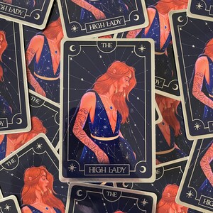 May include: A tarot card with a gold border and a blue background. The card features a woman with red hair and a blue dress. The text on the card reads "The High Lady".