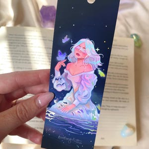 Può includere: Un segnalibro rettangolare con sfondo blu scuro raffigurante un cielo notturno, con l'illustrazione di una persona dai capelli e abiti iridescenti, circondata da farfalle e stelle luminose, con un cordino bianco in cima.