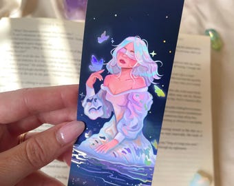Peut inclure: Un marque-page rectangulaire sur fond de ciel nocturne bleu foncé, illustrant une personne aux cheveux et vêtements iridescents, entourée de papillons et d'étoiles lumineuses, avec une boucle de cordon blanc au sommet.
