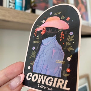 May include: A sticker featuring a cartoon illustration of a woman wearing a pink cowboy hat and a blue denim jacket. The background is black with floral accents. The text "COWGIRL Like me" is printed in white on the sticker.