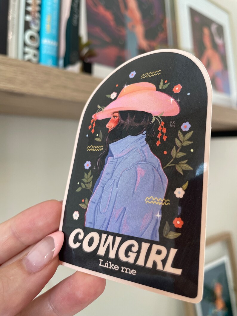 May include: A sticker featuring a cartoon illustration of a woman wearing a pink cowboy hat and a blue denim jacket. The background is black with floral accents. The text "COWGIRL Like me" is printed in white on the sticker.