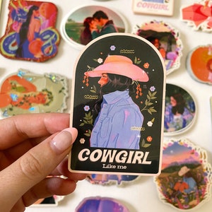 May include: A sticker with a black background and a pink and blue illustration of a woman wearing a cowboy hat. The text "COWGIRL Like me" is printed in white on a pink background.