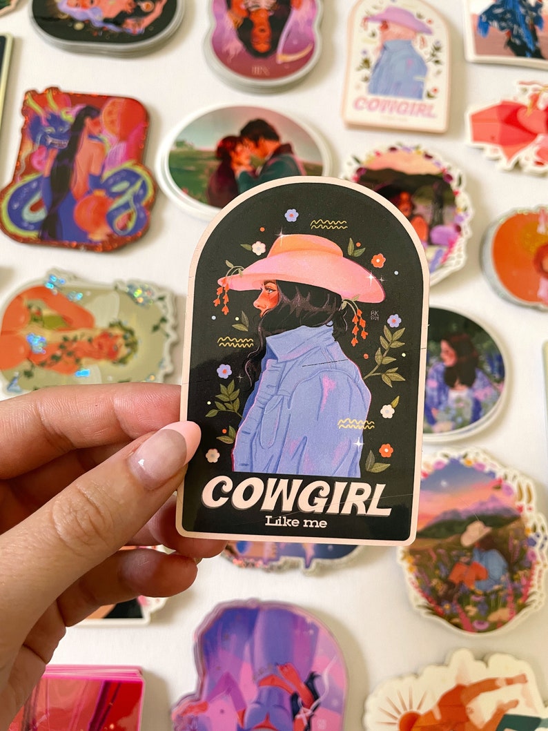 May include: A sticker with a black background and a pink and blue illustration of a woman wearing a cowboy hat. The text "COWGIRL Like me" is printed in white on a pink background.