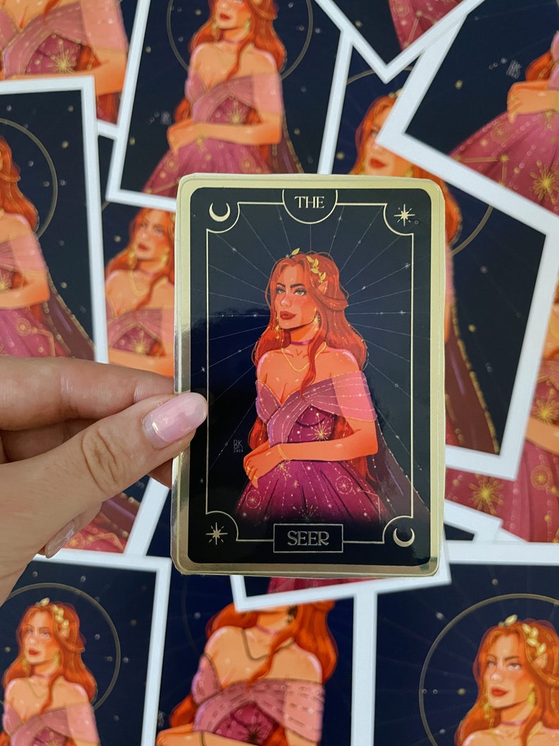 Op de afbeelding: Een tarotkaart met een gouden rand en een donkerblauwe achtergrond met een halve maan- en stervormig ontwerp. De kaart toont een vrouw met lang rood haar die een paarse jurk draagt met gouden accenten. De tekst "THE SEER" is op de kaart gedrukt.