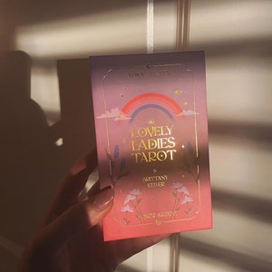 May include: The Lovely Ladies Tarot deck box features a pink and purple background with a rainbow, stars, and flowers. The text on the box reads "The Lovely Ladies Tarot" by Brittany Kehrer.