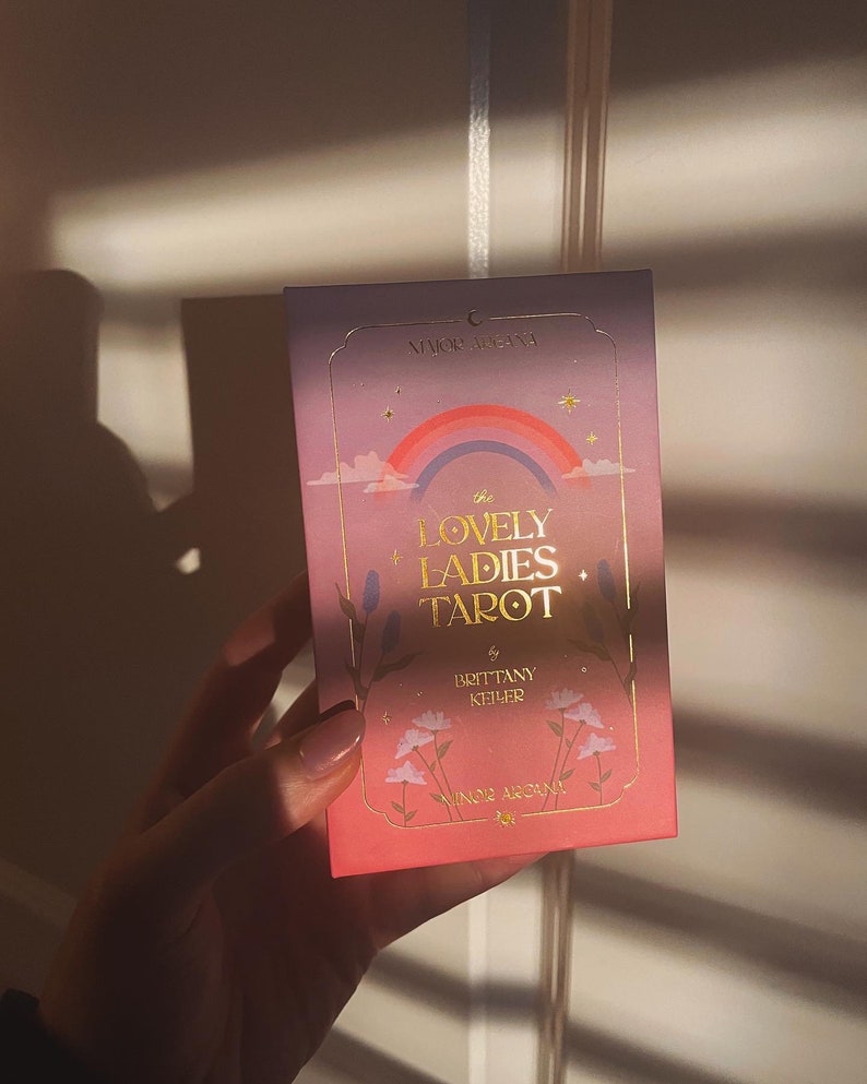 May include: The Lovely Ladies Tarot deck box features a pink and purple background with a rainbow, stars, and flowers. The text on the box reads "The Lovely Ladies Tarot" by Brittany Kehrer.