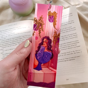May include: A pink and purple bookmark with a woman in a blue jacket looking out a window. The bookmark has a pink and purple background with hanging plants and a pink and purple sky. The bookmark has the text "BK 2023" in the bottom right corner.