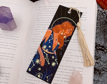 Op de afbeelding: Een zwarte bladwijzer met een gouden rand toont een vrouw met rood haar die een zwarte jurk met een sterrenbeeldpatroon draagt. De vrouw houdt een gloeiende bol in haar hand. De bladwijzer ligt op een boek met de tekst "A Court of Mist and Fury" en een roze kristal.