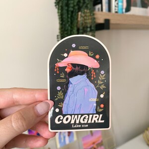 May include: A sticker featuring a cartoon illustration of a woman wearing a pink cowboy hat and a blue denim shirt. The sticker has a black background with white flowers and the text "COWGIRL Like me".