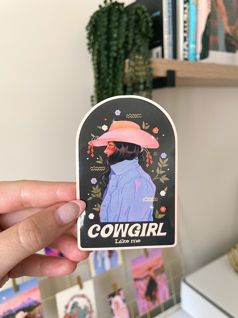 May include: A sticker featuring a cartoon illustration of a woman wearing a pink cowboy hat and a blue denim shirt. The sticker has a black background with white flowers and the text "COWGIRL Like me".