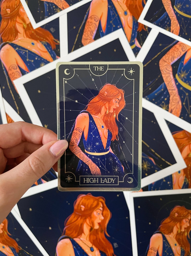 May include: A gold foil tarot card with a dark blue background and a woman with red hair wearing a blue dress with gold accents. The card reads "The High Lady" and has a crescent moon and a star in the corners.