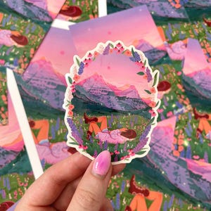 May include: A sticker featuring a mountain landscape scene with a person lying in a field of flowers. The sticker is surrounded by a floral border and has a pink and purple color scheme.