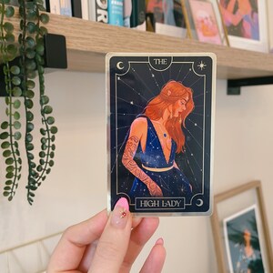 May include: A tarot card with a gold border and a blue background. The card features a woman with long red hair wearing a blue dress with gold stars. The card reads "THE HIGH LADY" in gold lettering.