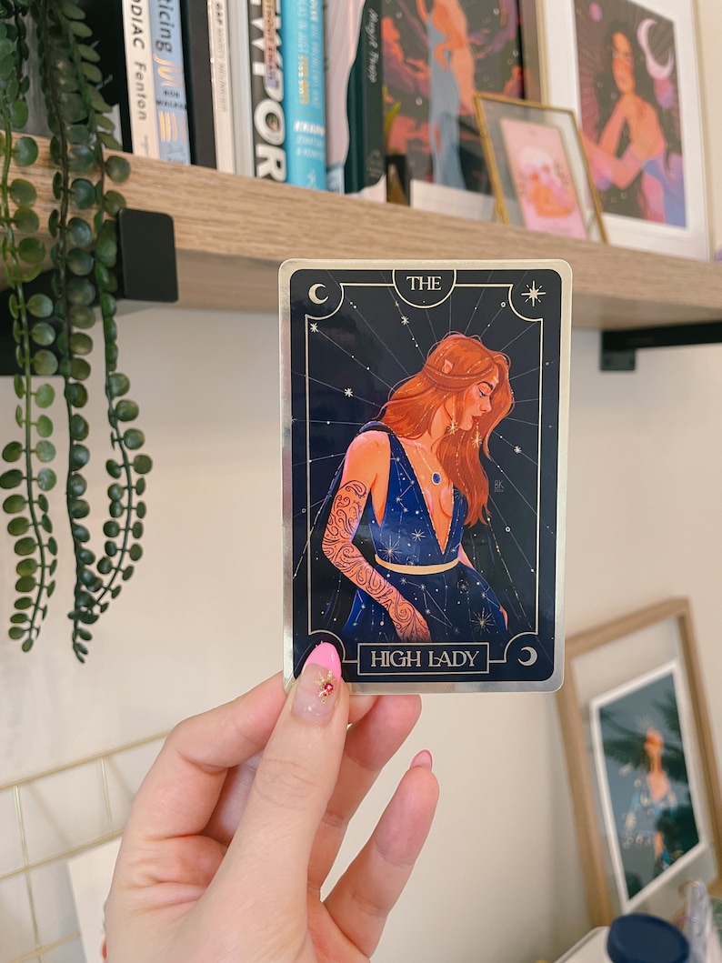 May include: A tarot card with a gold border and a blue background. The card features a woman with long red hair wearing a blue dress with gold stars. The card reads "THE HIGH LADY" in gold lettering.