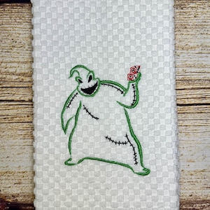 May include: White kitchen towel with an embroidered green and black image of the character Oogie Boogie from the movie The Nightmare Before Christmas. Oogie Boogie is holding a red bag of bugs in his right hand.
