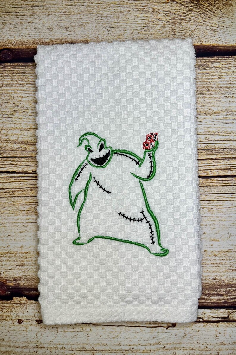 May include: White kitchen towel with an embroidered green and black image of the character Oogie Boogie from the movie The Nightmare Before Christmas. Oogie Boogie is holding a red bag of bugs in his right hand.