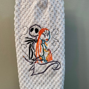 May include: White kitchen towel with an embroidered image of Jack Skellington and Sally from the Nightmare Before Christmas movie. The characters are in a heart shape with a swirl design.