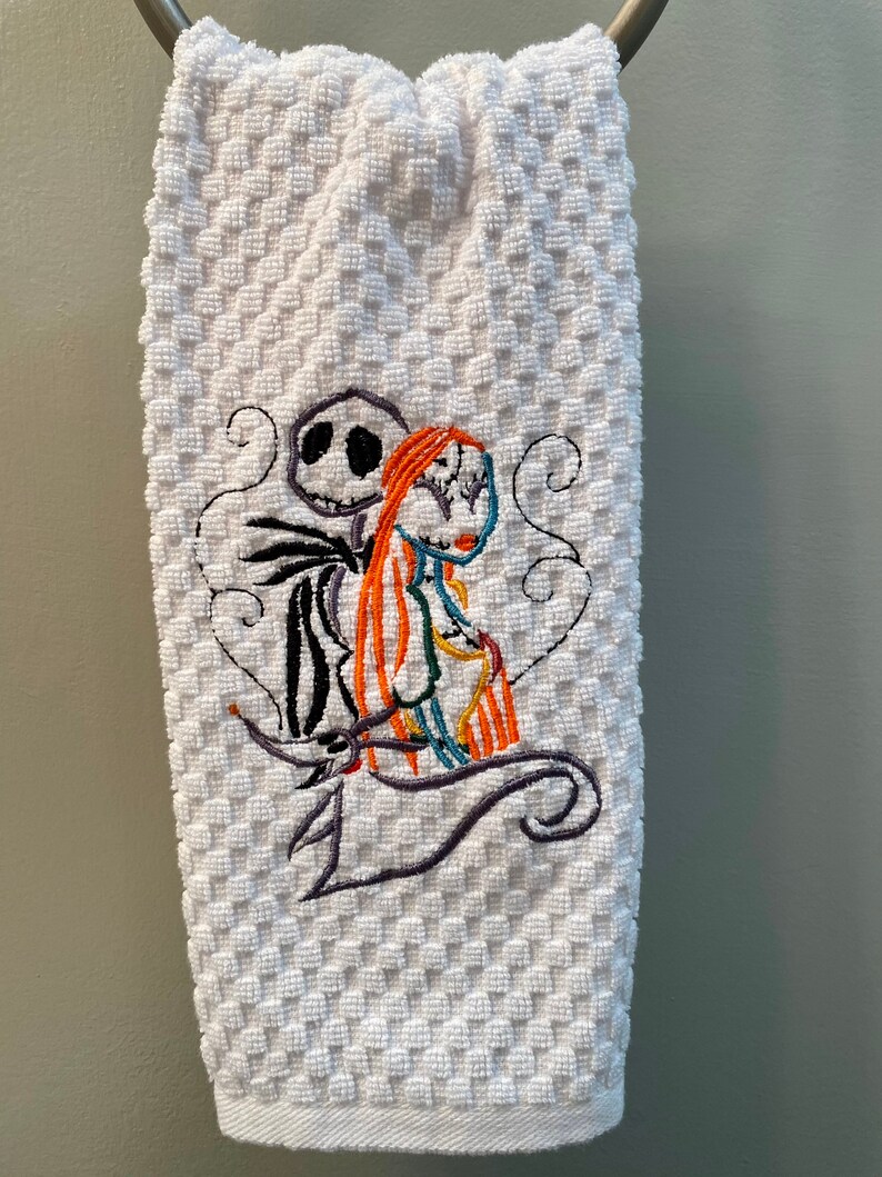 May include: White kitchen towel with an embroidered image of Jack Skellington and Sally from the Nightmare Before Christmas movie. The characters are in a heart shape with a swirl design.