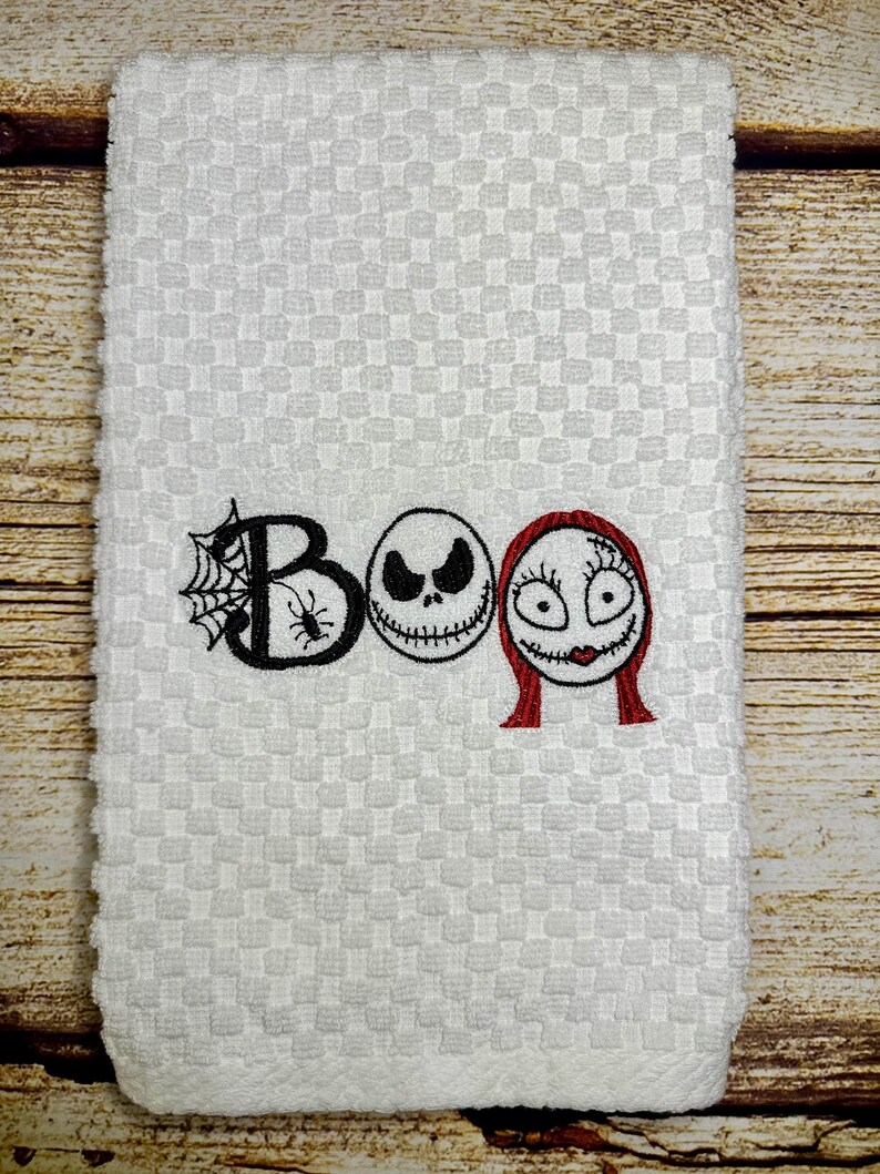 May include: White kitchen towel with an embroidered "BOO" design featuring the characters Jack Skellington and Sally from the movie The Nightmare Before Christmas.