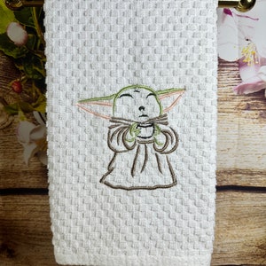 May include: White kitchen towel with an embroidered image of a green alien character with closed eyes holding a cup. The alien is wearing a brown robe.