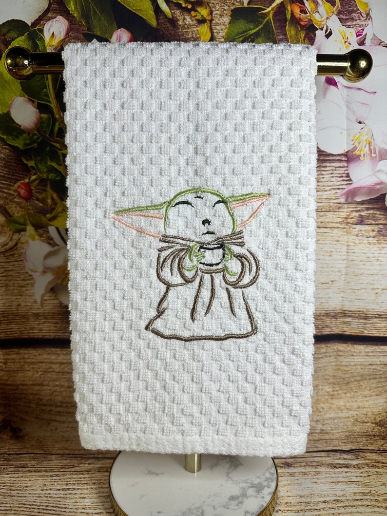 May include: White kitchen towel with an embroidered image of a green alien character with closed eyes holding a cup. The alien is wearing a brown robe.