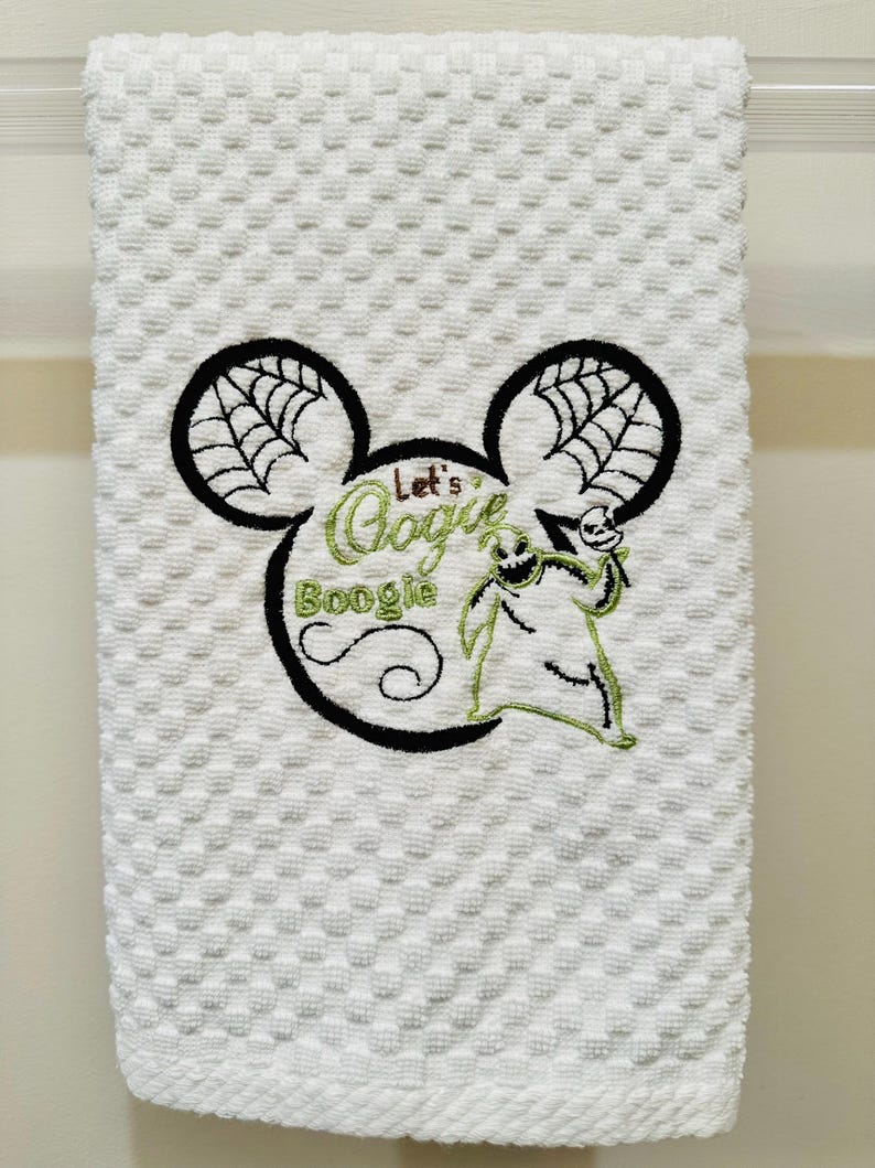 May include: White kitchen towel with an embroidered Mickey Mouse head design. The ears have a spiderweb pattern. The text "Let's Boogie" is inside the head with a green Oogie Boogie character.