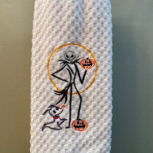 May include: White kitchen towel with an embroidered image of Jack Skellington from The Nightmare Before Christmas. Jack is holding a pumpkin and standing next to Zero, his ghost dog.
