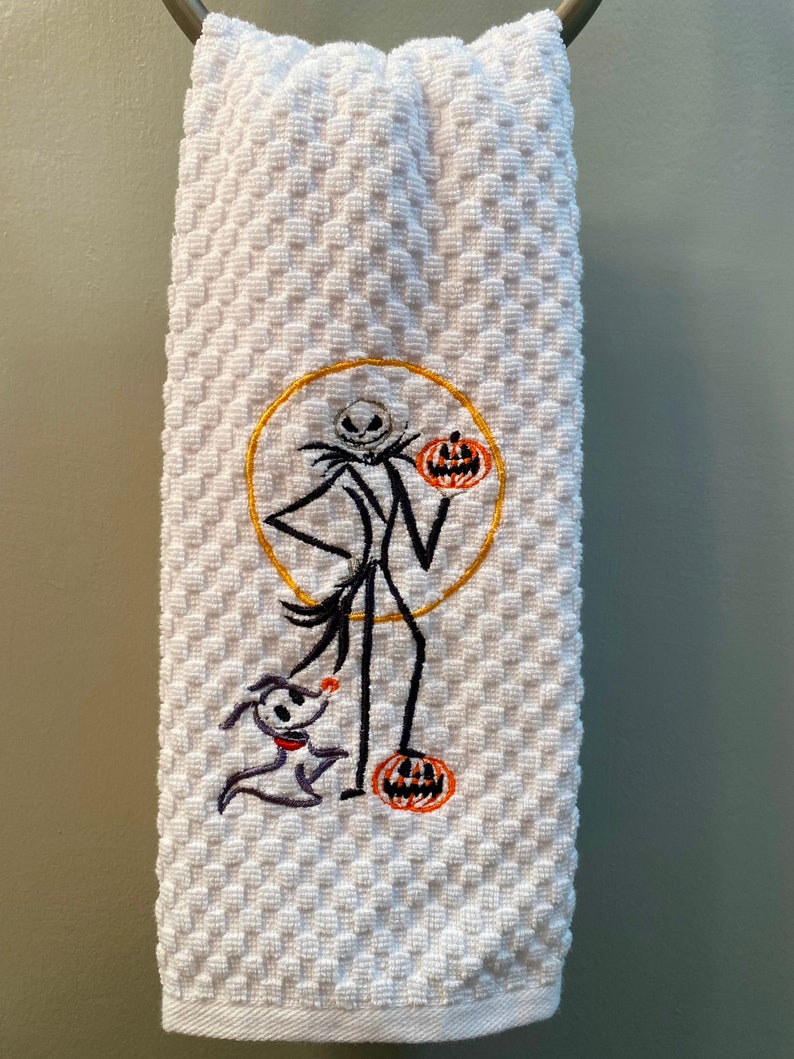 May include: White kitchen towel with an embroidered image of Jack Skellington from The Nightmare Before Christmas. Jack is holding a pumpkin and standing next to Zero, his ghost dog.
