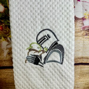 May include: White kitchen towel with an embroidered design of a Star Wars character holding a small green creature.