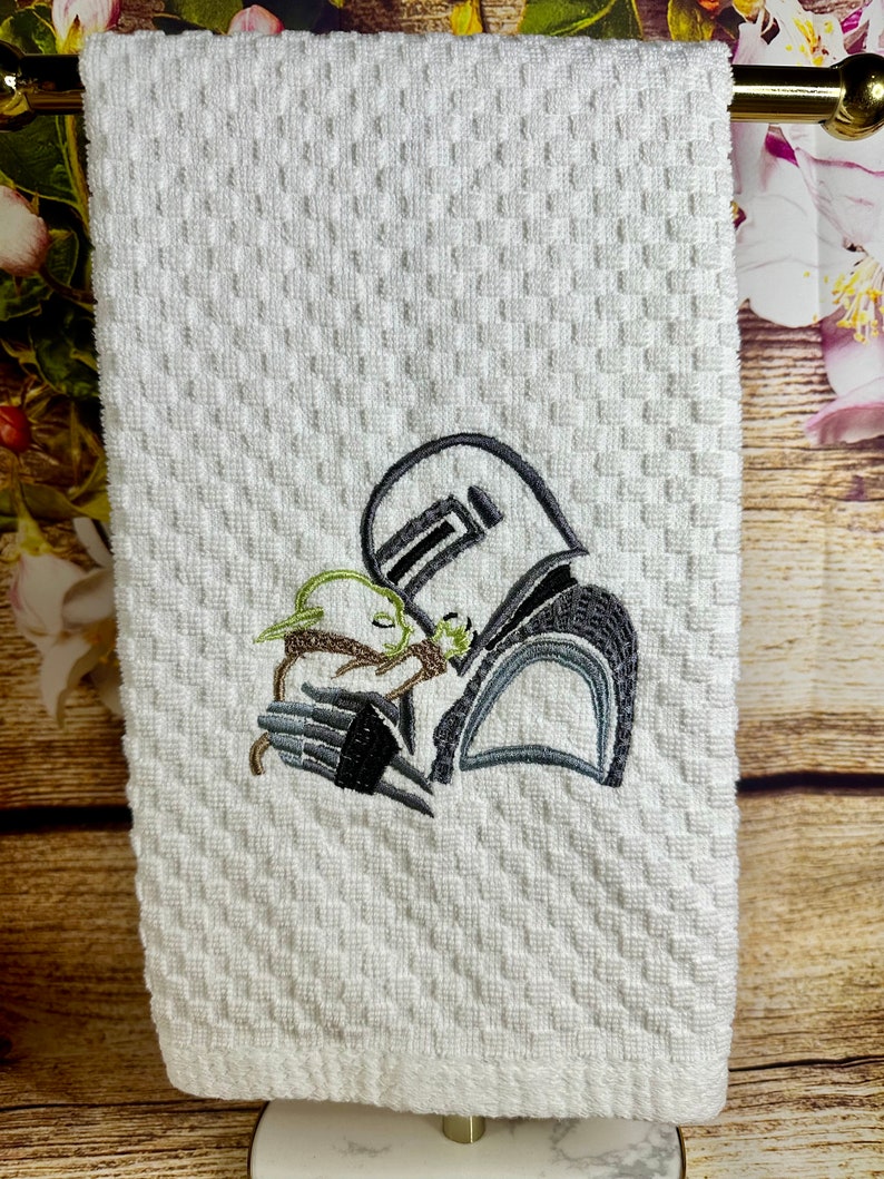 May include: White kitchen towel with an embroidered design of a Star Wars character holding a small green creature.