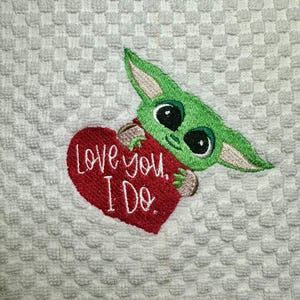 May include: A white kitchen towel with a green and red embroidered design. The design features a cartoon character holding a red heart with the text "Love you. I Do."